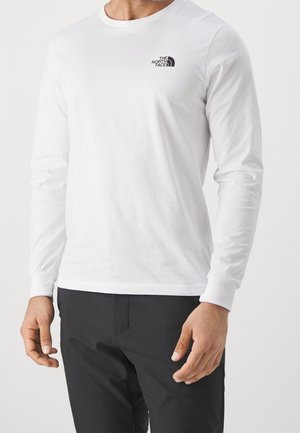 Mężczyzna w białej koszuli z długim rękawem z logo The North Face na piersi oraz czarnych spodniach, stojący z rozluźnionymi ramionami.