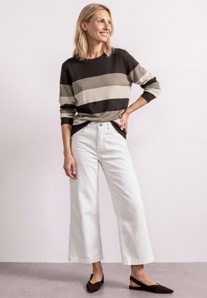 Vrouw lacht en kijkt zijwaarts, draagt een gestreept shirt met lange mouwen, witte wijde broekspijpen en zwarte puntige platte schoenen.