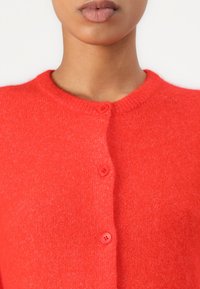 Red knitted cardigan, made of soft fabric, featuring a round neckline and five pink buttons down the front. Texture appears fuzzy.