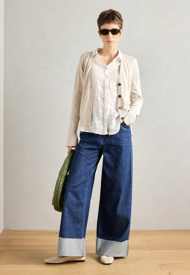 Modell trägt eine beigen Cardigan über einem weißen Hemd, weite blaue Jeans mit hellblauen Säumen und crèmefarbene Ballerinas, während sie eine grüne Tasche hält.