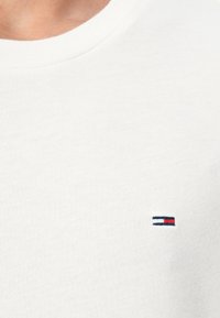 T-shirt en coton blanc avec un col rond et un petit logo brodé avec des bandes rouge, blanche et bleue sur la poitrine gauche.
