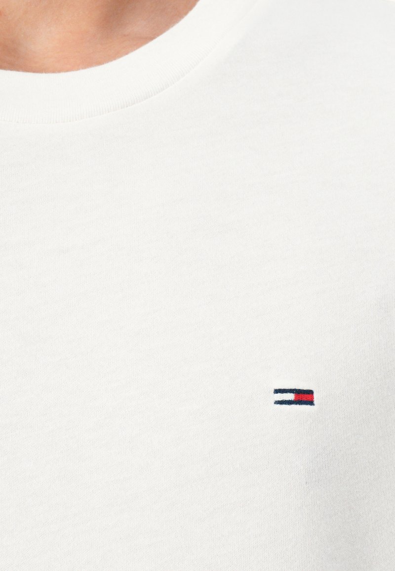 Camiseta de algodón blanca con un cuello redondo y un pequeño logo bordado con franjas rojas, blancas y azules en el lado izquierdo del pecho.