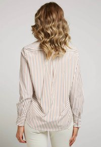 Chemise rayée en crème et marron. Dispose de manches longues, de poignets boutonnés, d'un dos cintré et d'une ourlet arrondi. Tissu lisse avec une légère brillance.