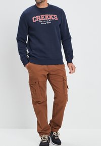 Sudadera de marina con gráfico "CREEKS" en rojo y blanco, combinada con pantalones cargo marrones que cuentan con múltiples bolsillos y un corte relajado.