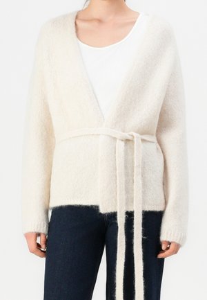 Femme portant un cardigan blanc doux et duveteux avec une ceinture nouée à la taille, par-dessus un top blanc et un jean bleu foncé.