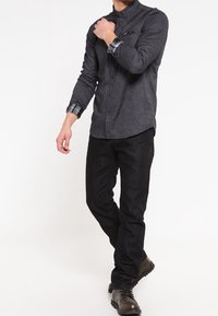 Mørkegrå skjorte med lange ærmer og ternede mansjetter, parret med sorte jeans og grønne støvler, stående foran en hvid baggrund.