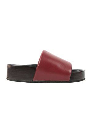 Sandalia de una sola pieza con suela negra gruesa y una ancha tira de cuero rojo en la parte superior, vista de lado sobre un fondo blanco.