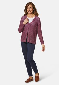 Violetter Strickcardigan mit offenen Maschen, langen Ärmeln, Knopfverschluss, kombiniert mit dunkelblauen Hosen und braunen Slippern. Darunter ein schlichtes weißes Oberteil.