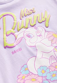 Hellviolettes Baumwoll-T-Shirt mit dem Schriftzug "Miss Bunny" in Grün und Rosa, versehen mit einer Grafik eines Kaninchens, das Blumen hält, akzentuiert durch Pastellfarben.