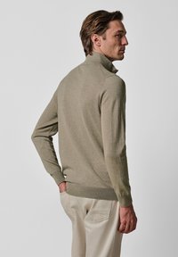 Jasnobeżowy sweter z dzianiny z kołnierzem stójką, długimi rękawami i ściągaczem u dołu. Noszony na jasnych spodniach. Gładka faktura.