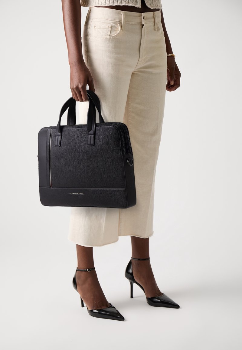 Personne portant un pantalon large crème et des talons noirs tenant une mallette noire en cuir à double poignée.