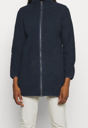 Vrouw die een marineblauwe fleecejas met ritssluiting en lange mouwen draagt, gecombineerd met lichtgekleurde broek, tegen een effen achtergrond.