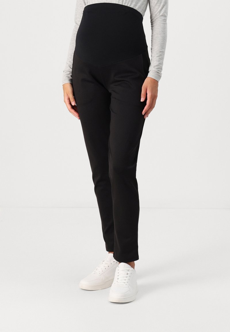 Personne enceinte portant un pantalon de maternité noir avec une ceinture élastique haute, un haut gris à manches longues et des baskets blanches, debout sur un sol blanc.