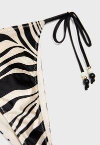 Dámske plavkové nohavičky so zebrovým pruhovaním, čiernymi bočnými šnúrkami ozdobenými čiernymi, bielymi a zlatými korálkami na svetlom pozadí.