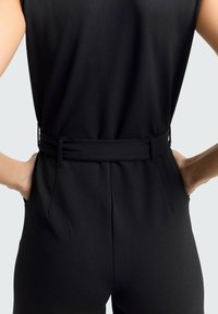 Combinaison noire avec une taille ajustée, détail ceinturé et poches latérales. Le tissu est lisse et mat, mettant en valeur un design épuré et ajusté.