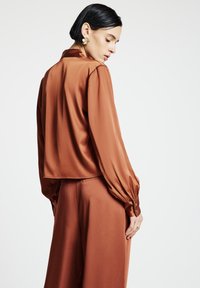 Woman with short dark hair wearing rust-colored satin blouse and matching wide-leg pants, facing sideways against plain white background.