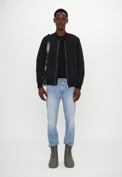 Veste bomber noire, chemise noire, jeans bleu clair, bottines vertes à zippers, se tenant contre un fond blanc.