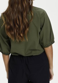 Lang golvend bruin haar valt over een olijfgroene top met pofmouwen en zwarte hoge taillebroek van een persoon die van zich af kijkt.