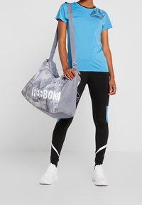 Grå duffelväska med Reebok-logotyp, texturerat mönster och justerbar rem; ihop med en blå T-shirt med kort ärm och svarta leggings.