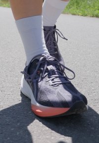 Chaussure de sport violette avec une tige en mesh texturé, des accents noirs et une semelle intermédiaire rose. Portée avec des chaussettes blanches sur un chemin gris.