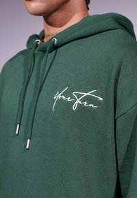 Sweatshirt à capuche vert en tissu doux, avec le texte en lettres cursives blanches "À Votre Tour" sur la poitrine et des cordons de serrage avec des embouts en argent.