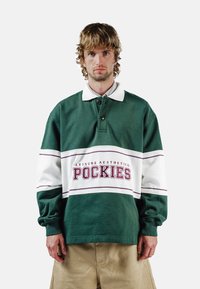 Grønn og hvit langermet poloskjorte med krage, med et dristig rødt grafisk trykk "POCKIES" og horisontale striper over brystet.