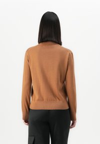 Vrouw met steil zwart haar, gekleed in een lange bruine trui met lange mouwen en zwarte broek, gezien van achteren tegen een witte achtergrond.