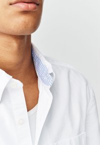 Chemise à col blanc avec col intérieur à rayures bleues. Tissu doux avec des boutons sur le devant et une texture subtile. Superposée à un t-shirt blanc.