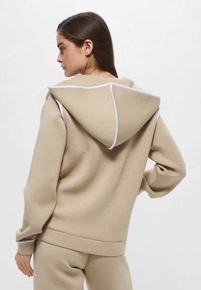 Felpa beige realizzata in tessuto morbido con un grande cappuccio e dettagli bianchi. Presenta polsini e orlo a coste, con un design dalla vestibilità morbida.
