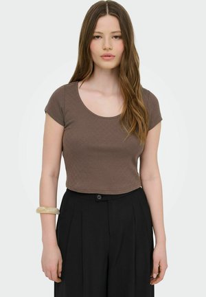 Jonge vrouw met lang bruin haar, gekleed in een nauwsluitend bruin kort mouwtopje, zwarte hoge taillebroek en een lichtkleurige armband aan haar linkerpols.