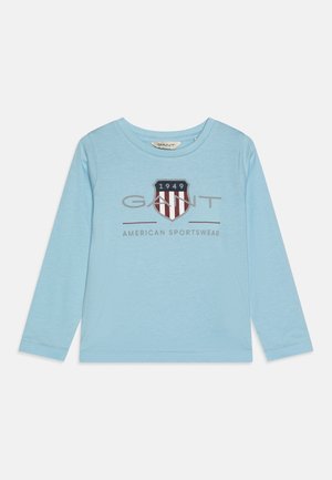 Heled sinise pika varrukaga T-särk, mis on valmistatud pehmest kangast ja millel on tekst "GANT AMERICAN SPORTSWEAR" ning kilbi logo punaste ja laevastiku värvi triipudega.