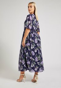 Mekko, jossa on kukkakuvio tummansinisellä taustalla, jossa on liila- ja vihreitä kukkia. Lyhyet hihat, vyötäröltä kavenettu ja hulmuavahelma. Kultaiset korkokengät.