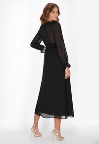 Schwarzes Maxikleid mit durchsichtigen, langen Ärmeln, gerafftem Taillenbund und feinen, strukturierten Punkten. Kombiniert mit goldenen High-Heel-Sandalen.