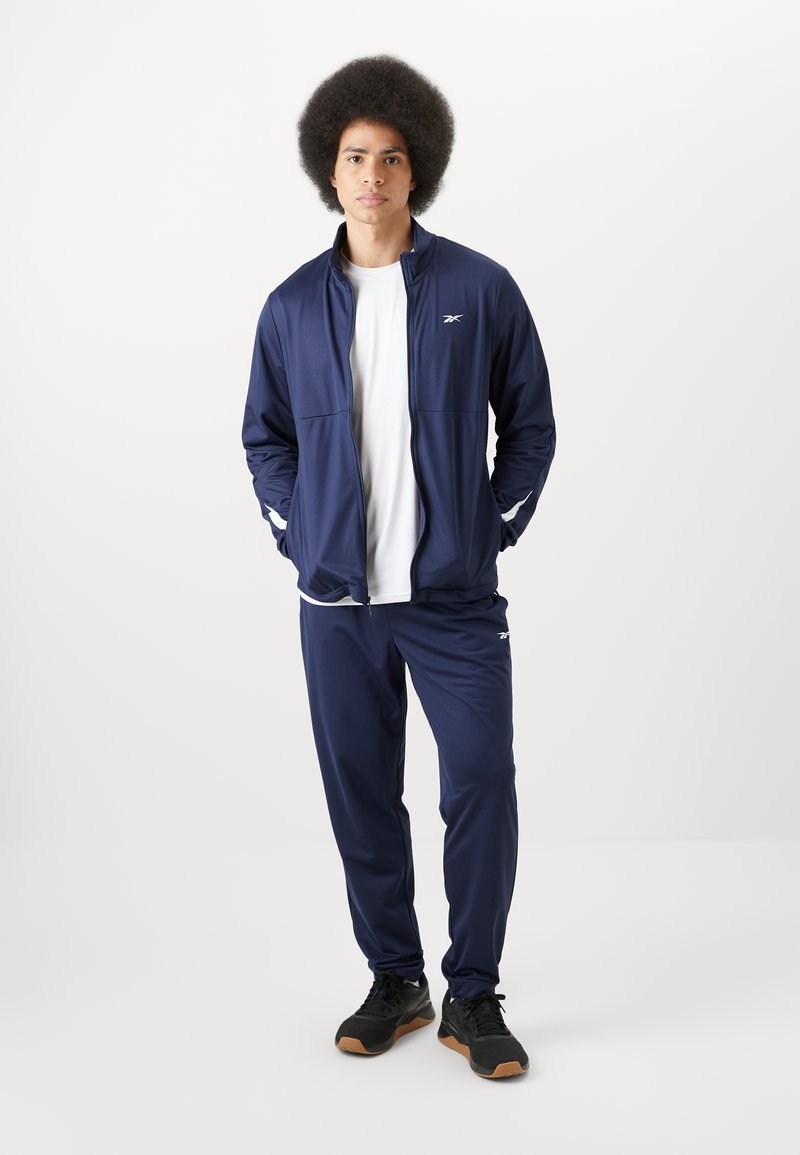 Chándal atlético de color navy hecho de tela suave con acentos en blanco. La chaqueta tiene cremallera completa y bolsillos laterales; los pantalones son ajustados con una cinturilla elástica.