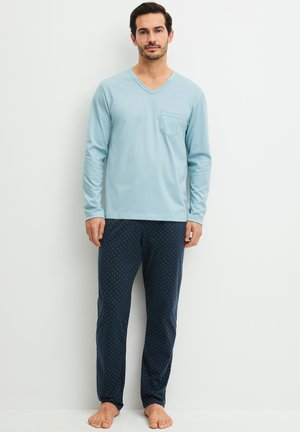 Uomo in piedi a piedi nudi che indossa una camicia azzurro chiaro a maniche lunghe e pantaloni da pigiama blu scuro con piccoli pois bianchi su uno sfondo semplice.
