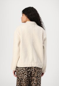 Cremefarbene Fleecejacke mit hohem Kragen und abfallenden Schultern. Weiche Textur, ausgestattet mit geradem Saum und minimaler Nahtdetails.