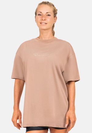 T-shirt oversize in cotone marrone chiaro, con maniche corte, scollo rotondo e piccolo dettaglio testuale bianco sul petto.