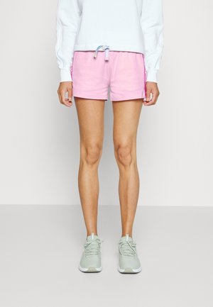 Vrouw in lichtroze trekshorts, een witte sweatshirt en lichtgroene sneakers, staande tegen een effen achtergrond.