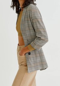 Marynarka w kratę w odcieniach szarości, brązu i bieli z jednym guzikiem. Nosi się ją na brązowej bluzce crop top, w połączeniu z beżowymi spodniami.