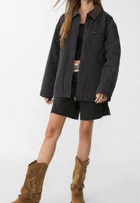 Persoon die een zwart oversized jack, zwart crop top, zwart rafelig korte broekje, cowboylaarzen en een zilveren riem draagt, met meerdere ringen aan de linkerhand.