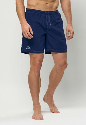 Mand iført mørkeblå badeshorts med hvide syninger og et lille logo på venstre ben, stående barfodet mod en ensfarvet baggrund.