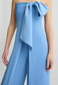 Blauer Jumpsuit aus leichtem Stoff mit einer großen Schleifenakzent am Ausschnitt und weitem Bein-Design, glatter Textur und tailliertem Schnitt.