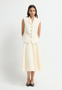 Elfenbensfarvet ærmeløs blazer med guldknapper og to forlommer, stylet med en matchende midi-nederdel. Modellen har sorte hæle på.