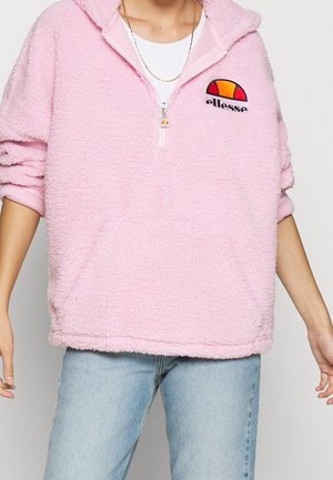 Person trägt einen hellrosa flauschigen Hoodie mit einem halben Reißverschluss und einem orange-roten ellesse-Logo auf der linken Brust, kombiniert mit hellblauen Jeans.