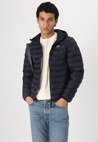 Marineblauw pufferjack met een ritssluiting aan de voorkant, capuchon en geribde manchetten. Heeft een klein groen logo en is gestyled over een wit shirt en blauwe jeans.