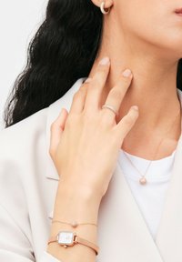 Orologio oro rosa con bracciale in rete, braccialetto con charm scintillante, anello con pietre trasparenti e collana con pendente circolare su abbigliamento chiaro.