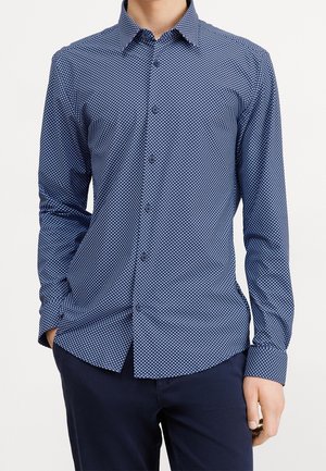 Homme portant une chemise bleu marine ajustée à manches longues avec de petits motifs géométriques blancs et un pantalon bleu marine foncé.