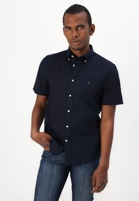 Camicia a maniche corte blu navy con colletto, bottoni bianchi e un piccolo logo sul petto; indossata con jeans in denim blu.