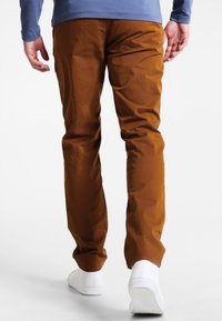 Pantaloni in cotone marrone con vestibilità slim, caratterizzati da una texture liscia, abbinati a scarpe bianche, visti da dietro.