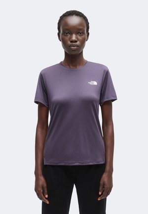Mujer con una camiseta morada lisa de North Face y pantalones negros, de pie frente a un fondo gris claro, mirando hacia adelante.